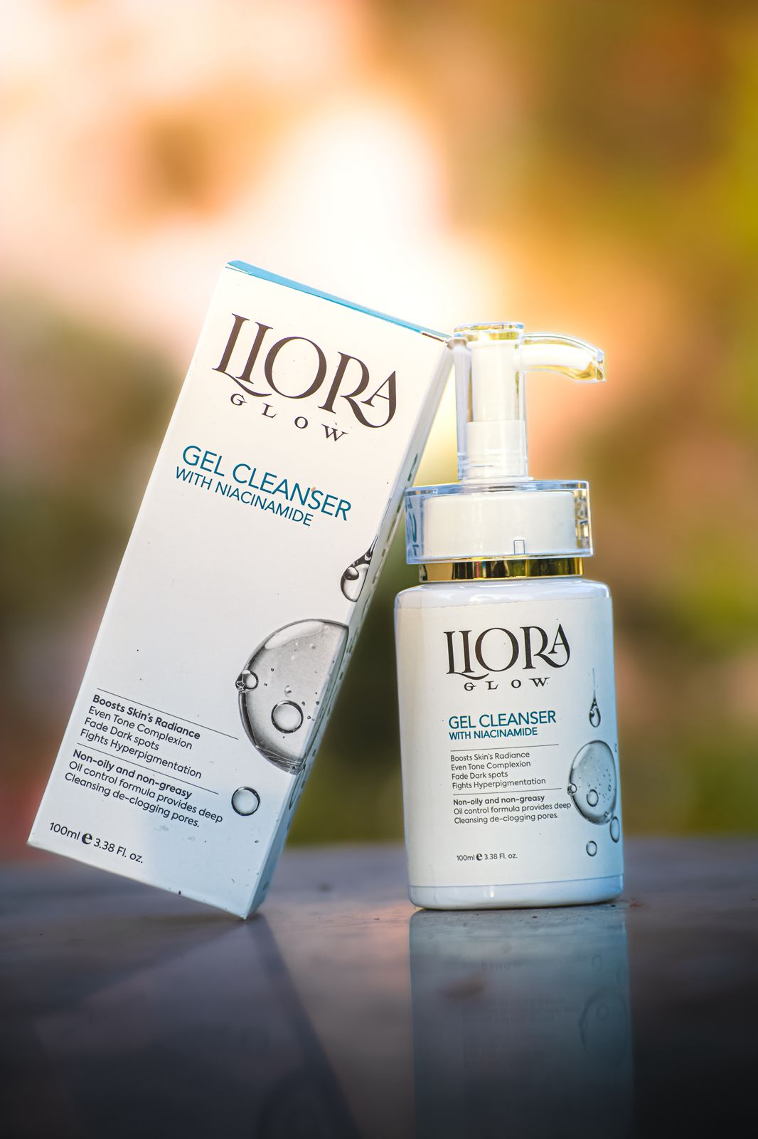 LLORA GLOW gel cleanser with pump and packaging on a reflective surface with blurred natural background